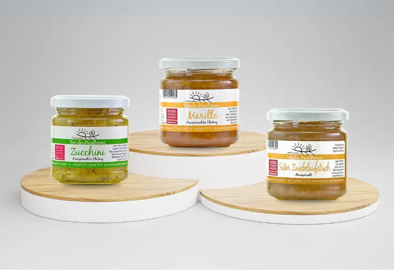 Drei Gläser hausgemachter Produkte vom Familienbetrieb Haselberger im Weinviertel: Zucchini-Chutney, Marillen-Aufstrich und Süßer Zwiebelaufstrich. Die Gläser stehen auf runden Holzpodesten.