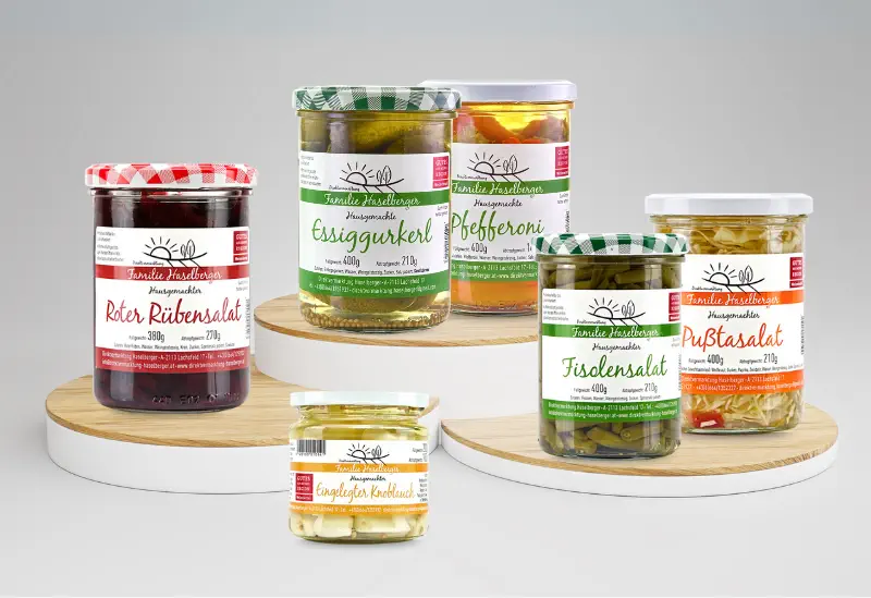 Verschiedene Gläser mit hausgemachten eingelegten Produkten vom Familienbetrieb Haselberger im Weinviertel. Zu sehen sind Roter Rübensalat, Essiggurkerl, Pfefferoni, Fisolen, Pußtasalat und eingelegter Knoblauch, dekorativ auf Holzpodesten präsentiert.